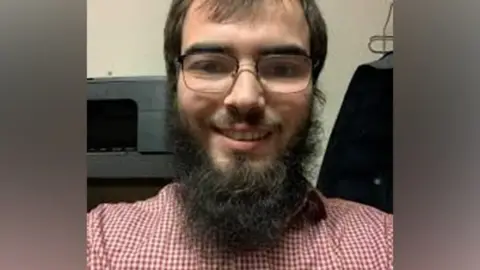 Met Police Khaled Hussein has brown hair, a large beard and glasses and a red and white shirt - the picture appears to be a selfie and he is smiling into the camera dqxikeidqkikdinv