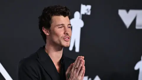 PA Media Shawn Mendes who has dark fairly short wavy hair and stubble. He is wearing a black jacket and looking towards his left with his hands up in prayer dqxikeidqkikdinv
