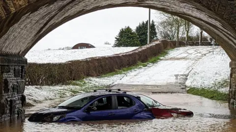 Supplied Two cars - one blue and one red - stuck in flooding under a bridge. Snowy fields and a road can be seen in the background. dqxikeidqkikdinv
