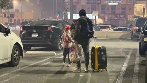Christal Hayes A woman with her daughter stands on a road with a suitcase in one hand and a bag in another. dqxikeidqkikdinv
