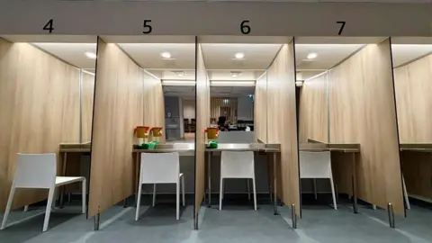 Numbered booths with a white chair in each booth. Each one faces a mirrored wall. It is brightly lit. dqxikeidqkikdinv