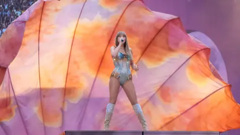 PA Media Taylor Swift on stage at a London concert, in front of a large peach and light purple coloured backdrop. She is singing into a microphone and wearing a one-piece and knee-high boots. dqxikeidqkikdinv