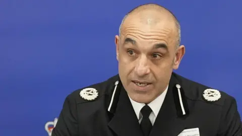 PA Media Thom has short cropped grey and black receding hair and brown eyes. He is wearing a black police uniform blazer with crest logo on both shoulders over a white shirt and black tie. He is talking while looking to the left. dqxikeidqkikdinv