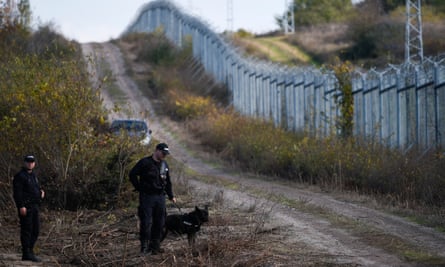 Two policemen in black uniforms, one with a guard dog on a leash, near a metal fence topped by barbed wire in countryside dqxikeidqkikdinv