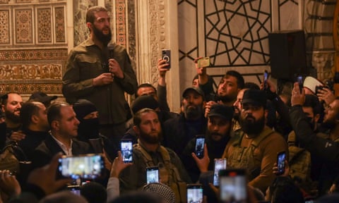 Sharaa speaks to a crowd of men at a mosque: most are wearing khaki shirts and military fatigues, and many in the audience are filming him dqxikeidqkikdinv