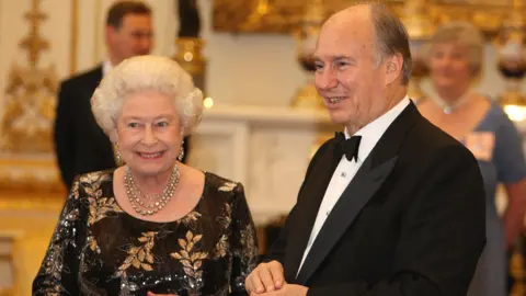 PA Media Queen Elizabeth II wearing a black dress with golden leaf patterns standing next to the Aga Khan wearing a black tuxedo dqxikeidqkikdinv