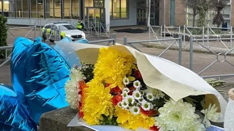 BBC/Simon Thake Flowers and balloons left on the wall outside the school building. A police car and officer can be seen in the background, near the entrance. dqxikeidqkikdinv