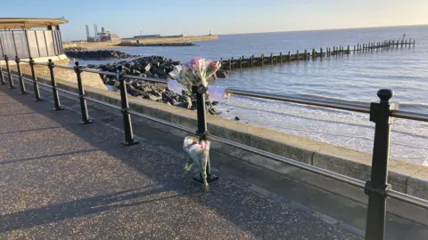 Luke Deal/BBC FLowers and tributes along a sea wall and barrier dqxikeidqkikdinv