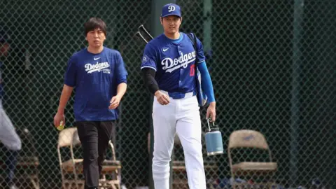 Getty Images Ippei Mizuhara, the former interpreter to Los Angeles Dodgers’s Shohei Ohtani, seen walking with the baseball star on the field. He is wearing a blue Dodgers shirt and Shohei is in his baseball uniform. dqxikeidqkikdinv