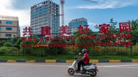Jonathan Head/ BBC A man riding past a red sign saying &rsquo;Let&rsquo;s build a beautiful home together&rsquo;  