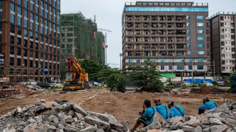 Jonathan Head/ BBC Workers in blue shirts sit amid construction rubble in front of tall buildings near a crane.
