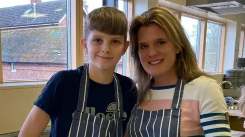 Ellen Roome A woman stands with a young boy. They are both wearing aprons and are smiling. dqxikeidqkikdinv