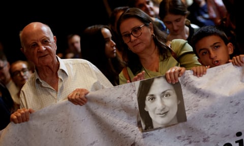 A man, woman and a boy hold a sheet with a b/w picture of Caruana Galizia on it dqxikeidqkikdinv