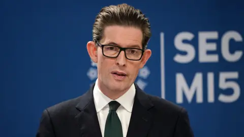 PA Media Ken McCallum, Director General of MI5 - a middle-aged man with slicked back dark brown hair, and wearing dark-rimmed rectangular glasses. He is wearing a dark suit, white shirt and navy blue tie. dqxikeidqkikdinv