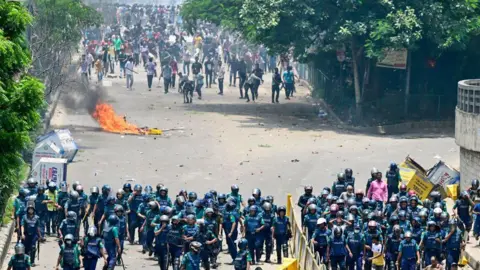Getty Images A group of police in blue uniforms and helmets, with their backs facing a crowd of protesters. A burning object lies on the ground between them. dqxikeidqkikdinv