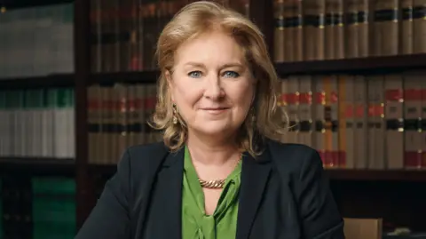 Rory Lindsay/UK Judiciary Baroness Sue Carr sits in front of a bookshelf with her arms crossed. She’s wearing a green shirt and black suit jacket. dqxikeidqkikdinv