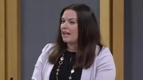Vikki Howells in the Senedd. She is stood at a lecturn about to speak. She is wearing a black top underneath a lilac jacket and has a beaded and gold necklace on. Her dark shoulder-length hair is untied. dqxikeidqkikdinv