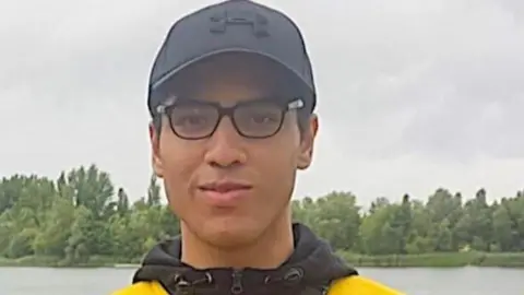 Facebook Nicholas Prosper dressed in glasses, a black baseball hat and yellow and black hoodie while standing in front of a lake. dqxikeidqkikdinv