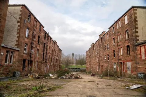 Sandstone fronted tenements. The windows are smashed and rubble is piled up in the street. dqxikeidqkikdinv
