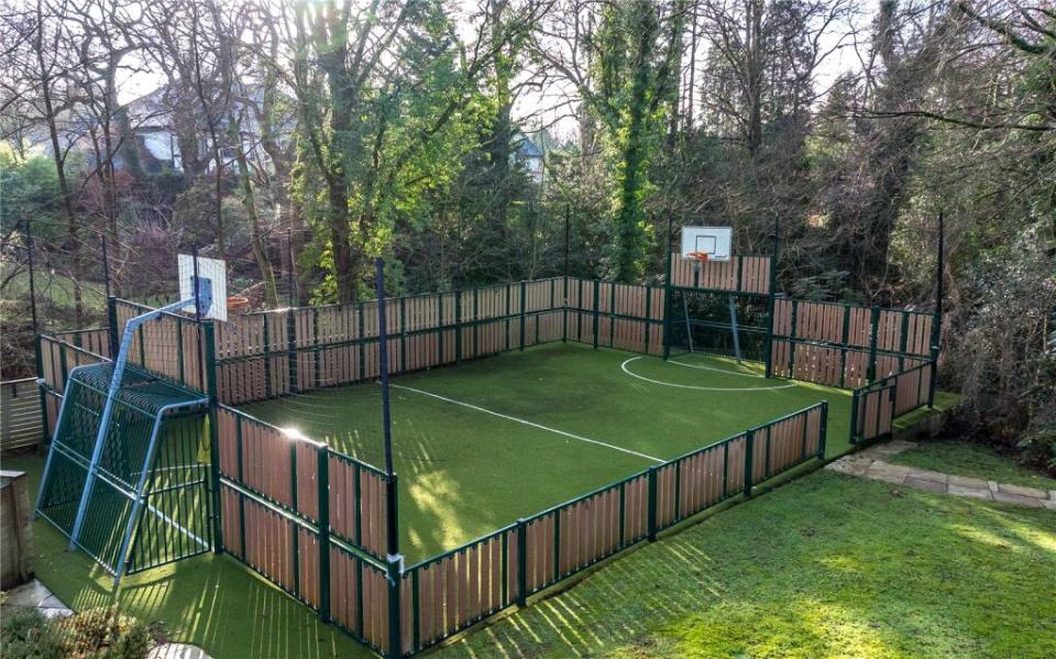 Multi-sport court with artificial turf, basketball hoops, and soccer goals, enclosed by a wooden fence.